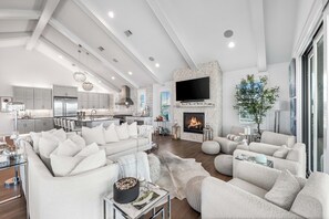 Ein modernes Wohnzimmer mit Kamin, großem Flachbildfernseher und Essbereich, im Hintergrund ist eine Küche zu sehen.