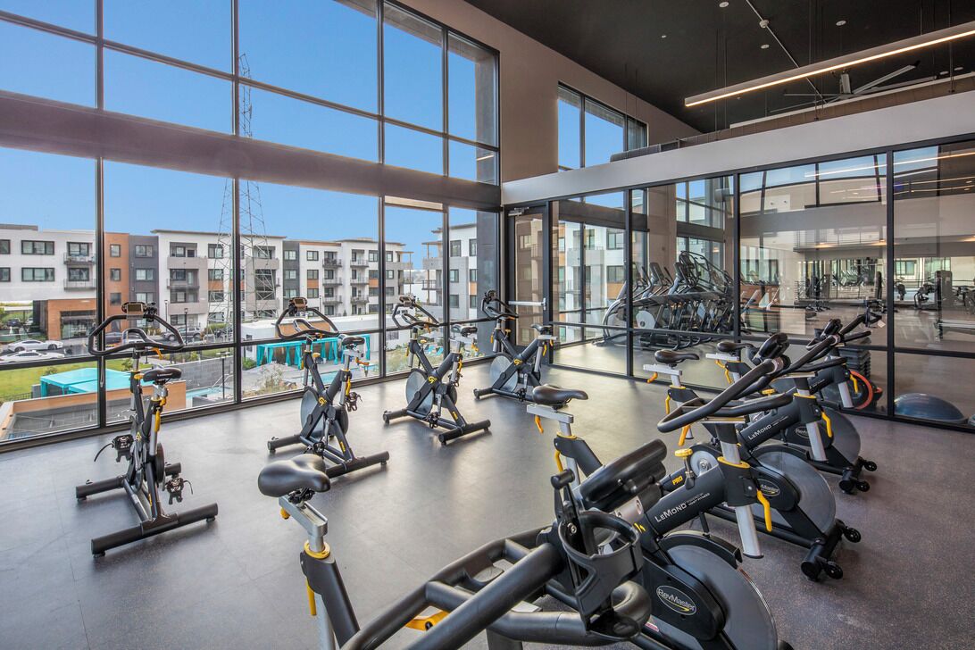 Un gimnasio moderno con varias bicicletas estáticas, grandes ventanales y vistas de edificios circundantes.