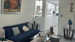 Un salón con un sofá azul oscuro, un sillón blanco, un ventilador de techo, un cuadro y vistas a través de una puerta abierta.