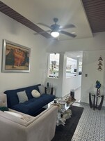 A living room with a dark blue sofa, a white armchair, a ceiling fan, a painting, and a view through an open door.