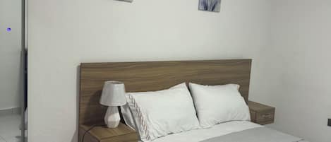 A bedroom with a wooden headboard, a bedside table with a lamp, a nightstand, and a wall-mounted air conditioner.