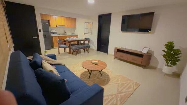 Ett vardagsrum med en soffa, ett soffbord, en tv, en matplats med stolar och ett kök i bakgrunden.
