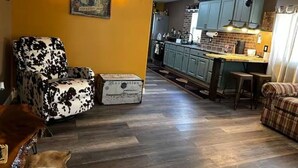 Una sala de estar con un sillón estampado, una mesita y un sofá. Se ve una cocina al fondo, con armarios azules y un revoque de ladrillo.
