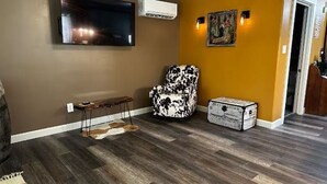 Un salón con televisor de pantalla plana, aire acondicionado, un sillón estampado, una mesita y un objeto decorativo.