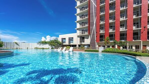 Una piscina con bordes azulejados, un edificio de varios pisos con balcones y un cielo azul despejado.