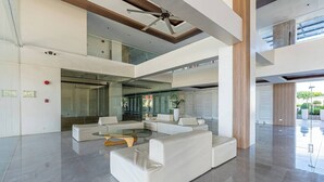Un vestíbulo amplio con diseño moderno, que incluye un área de descanso con sofás y una mesa de centro de cristal, un ascensor de vidrio y un techo alto con ventilador.