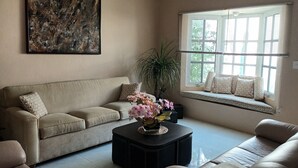 A living room with a beige sofa, a black coffee table with a flower vase, a large abstract painting, and a window with blinds. - La palma Great Location Spacious Funtonal Equipied (Cancún)