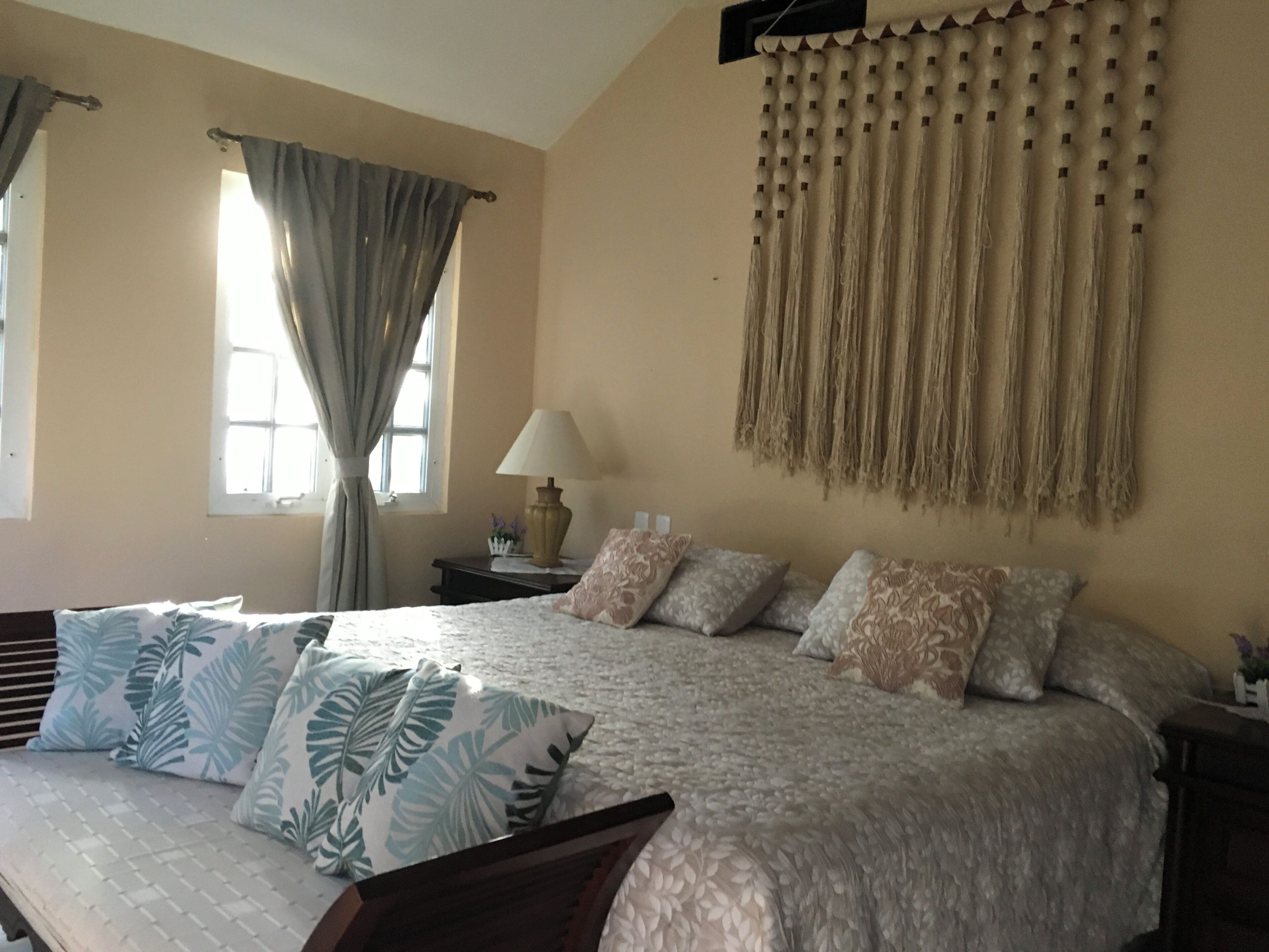 A bedroom with a bed, pillows, a nightstand, a lamp, a window with curtains, and a wall hanging.