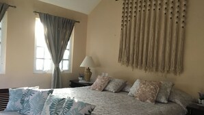 A bedroom with a bed, pillows, a nightstand, a lamp, a window with curtains, and a wall hanging. - La palma Great Location Spacious Funtonal Equipied (Cancún)