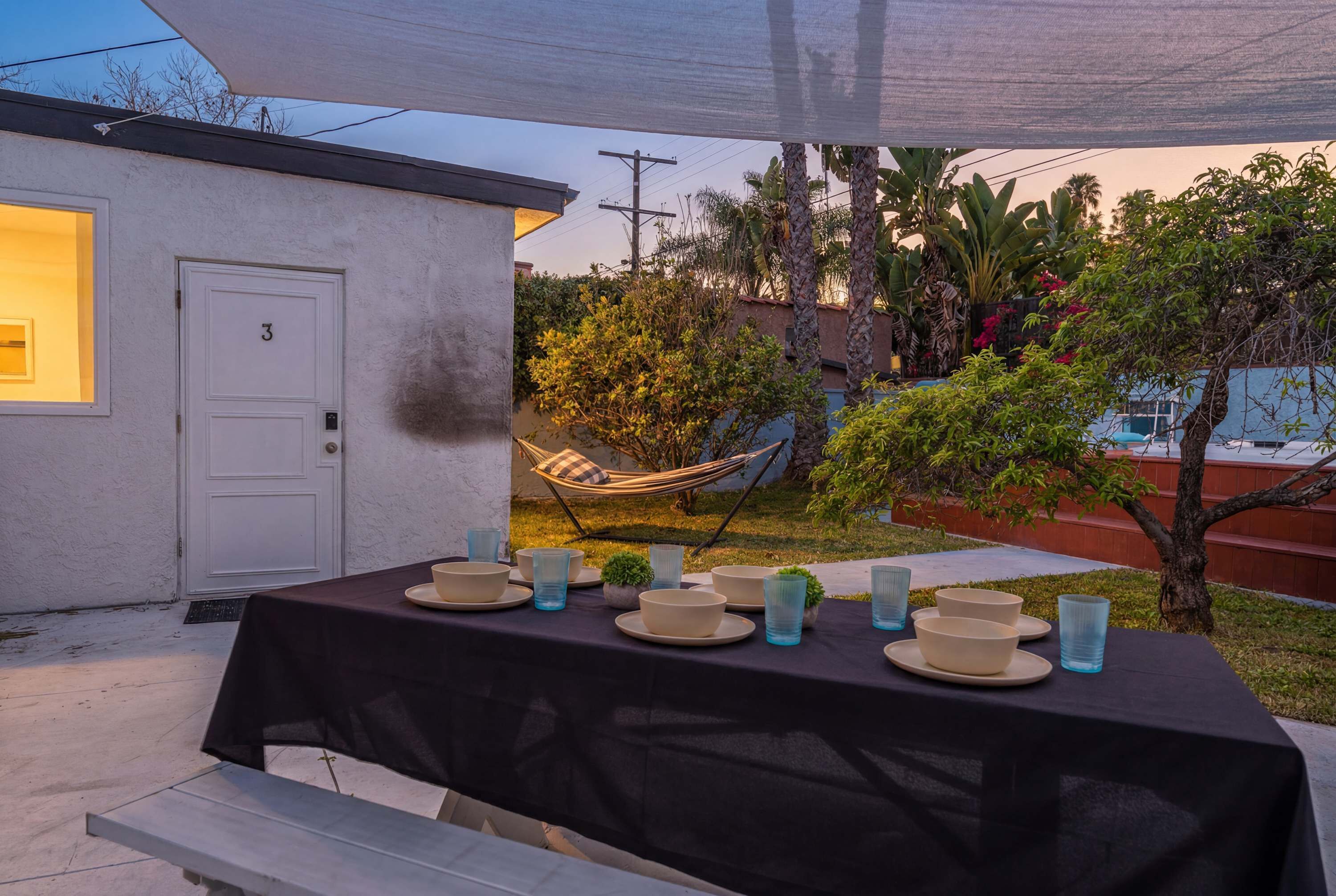 An outdoor dining area with a table set for four, surrounded by greenery and a hammock.