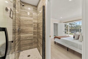 A modern bathroom with a glass shower enclosure and a bedroom with a bed and window.