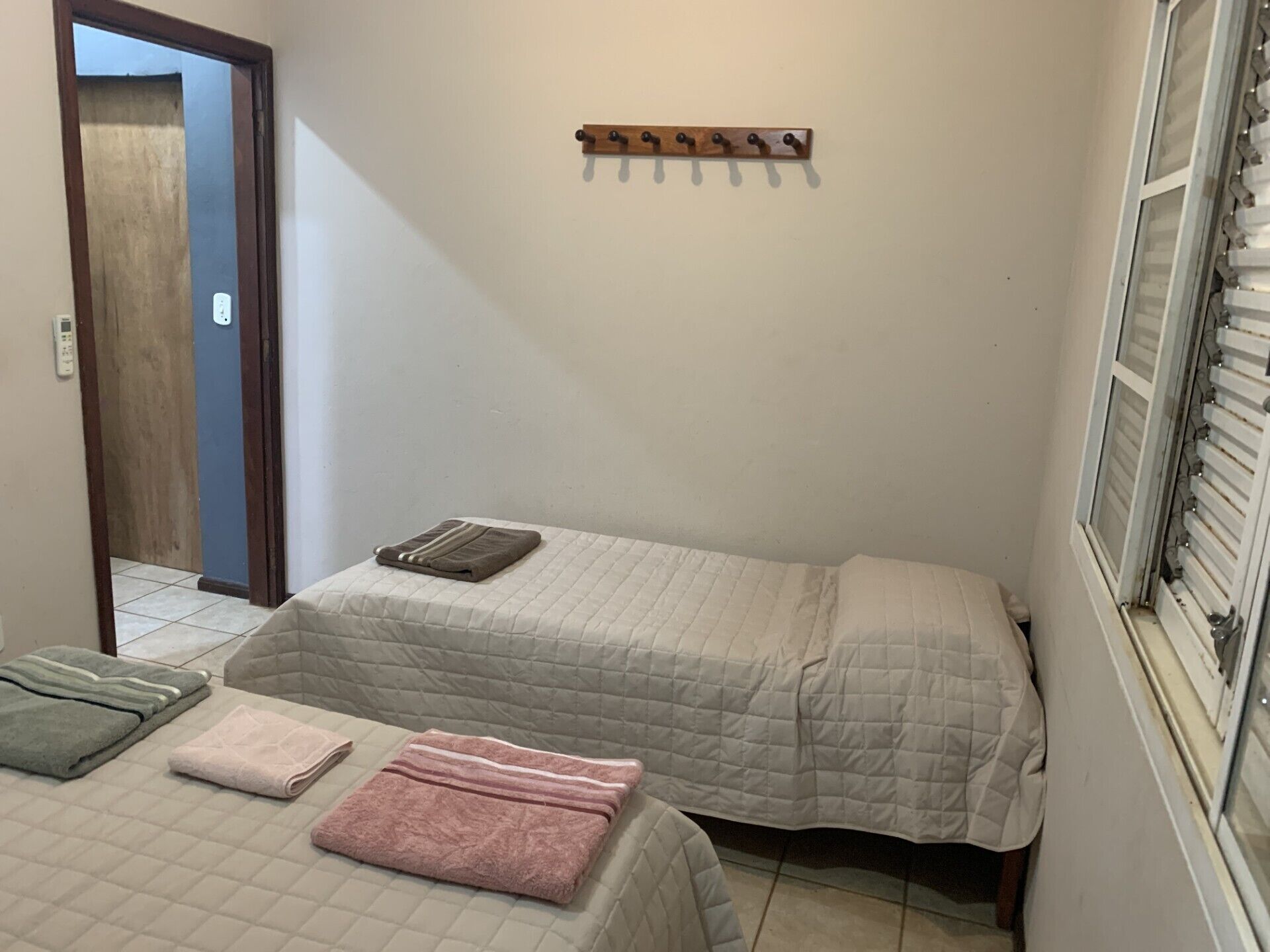 Una habitación pequeña y limpia con dos camas, un perchero de madera, una ventana con persianas y una puerta que da a otra habitación.