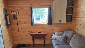 A cozy cabin interior with a wooden ceiling, a sofa, a small table, a window with curtains, and a cabinet. - Cozy 1-bedroom private cabin  with AC and heat. Minutes to Sacanadaga Lake (Broadalbin)