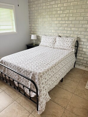 Un lit recouvert d’une couvre-lit à motifs floraux, une table de chevet avec une lampe et un mur dont le papier peint imite des briques.