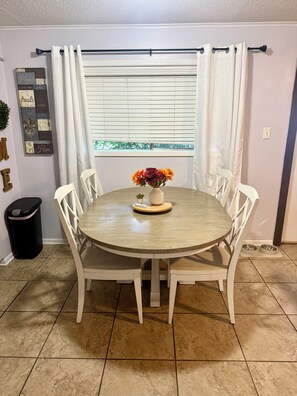 A dining area with a round table, white chairs, a vase with flowers, and a window with blinds.