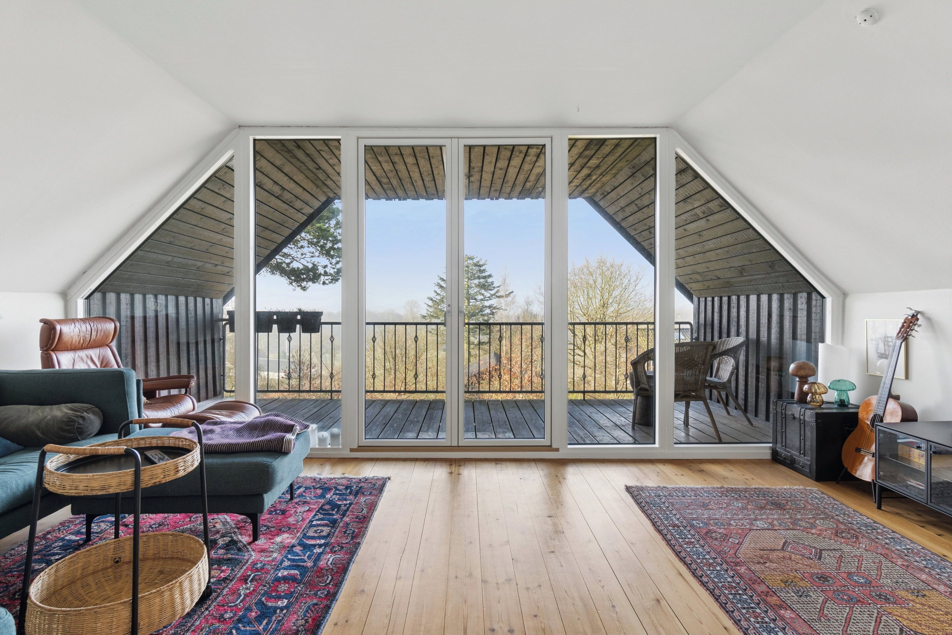 Une pièce spacieuse dotée d’une grande porte-fenêtre coulissante donnant sur une terrasse extérieure, d’un coin salon confortable avec un canapé et des fauteuils, et d’une guitare posée sur un support.