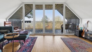 Un'ampia stanza con un'ampia porta-finestra che si apre su un patio esterno, un'area salotto accogliente con divano e poltrone, e una chitarra su un cavalletto.