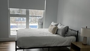 A bedroom with a bed, a nightstand, a window, and a view of the outside.