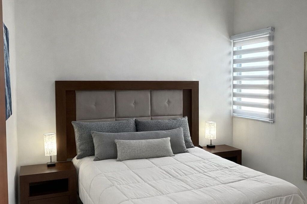 Camera da letto con letto, testiera, cuscini, comodini, lampade e finestra con persiane.