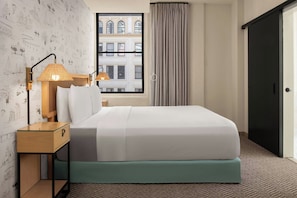 A hotel room with a bed, a nightstand, a lamp, a window with curtains, and a view of a building. - FOUND Hotels, San Diego Downtown, Series by Marriott  (San Diego)