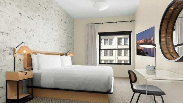 A hotel room with a bed, bedside table with lamp, chair, desk, large mirror, window with view, and patterned wallpaper. - FOUND Hotels, San Diego Downtown, Series by Marriott  (San Diego)