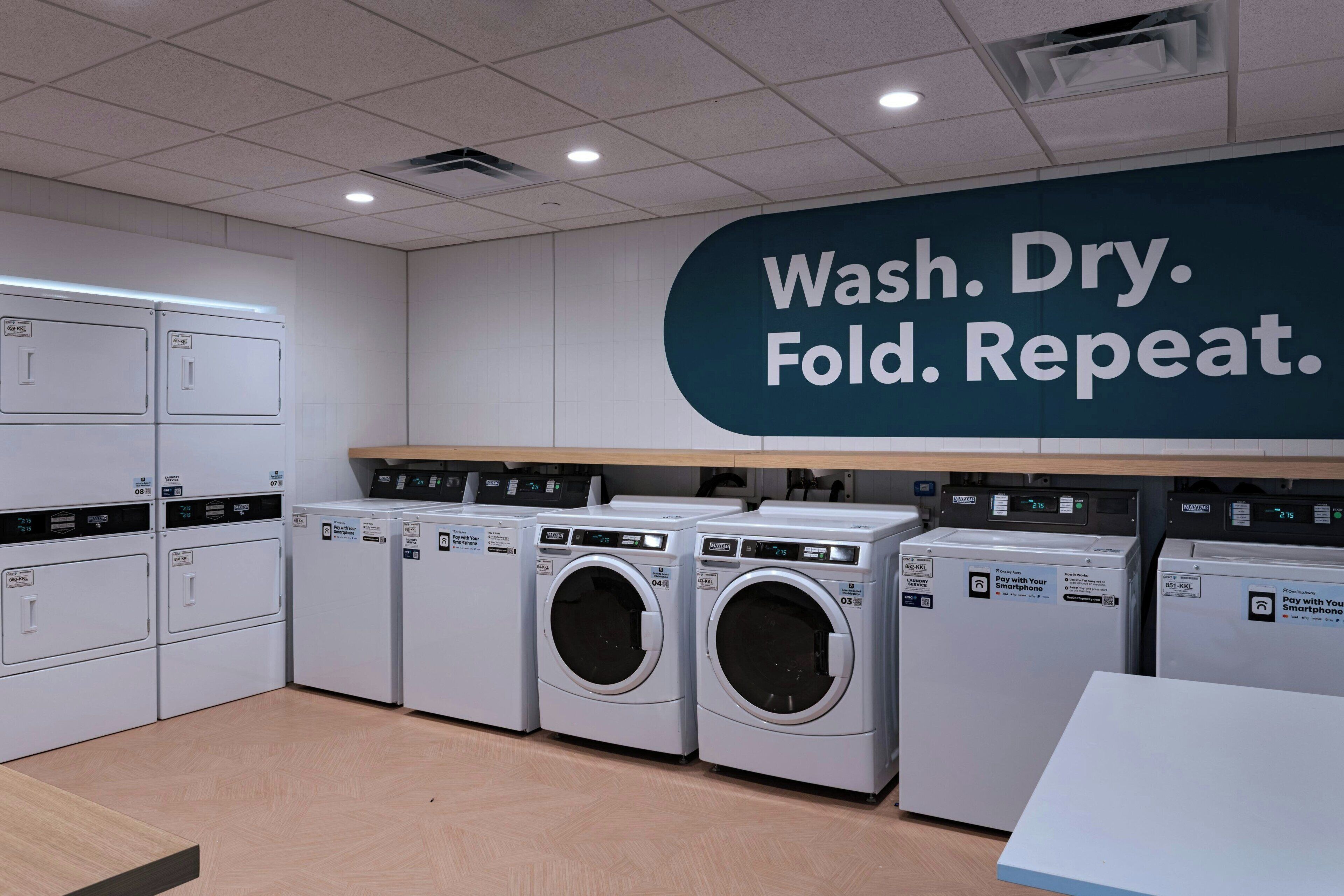 Una lavandería con varias lavadoras y secadoras, un cartel en la pared que dice 'Lavar. Secar. Doblar. Repetir.', y un terminal de pago para pagos móviles.