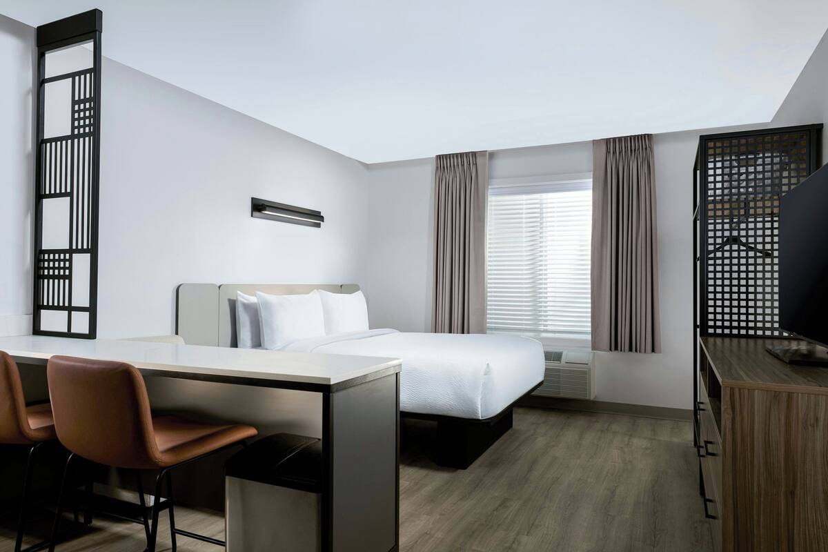 Une chambre d’hôtel comprenant un lit, un bureau avec des chaises, une télévision et une fenêtre avec des rideaux.