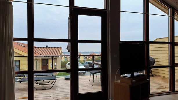 A room with large windows, a TV on a stand, and a view of a deck with outdoor furniture. - Holiday home near forest and beach (Egernsund)