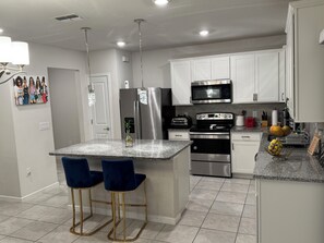 A modern kitchen with white cabinets, stainless steel appliances, a granite countertop, and two blue bar stools. - Modern 3BR Home w/ Open Floor Plan, Huge Yard & Security System – Move-In Ready (Ruskin)
