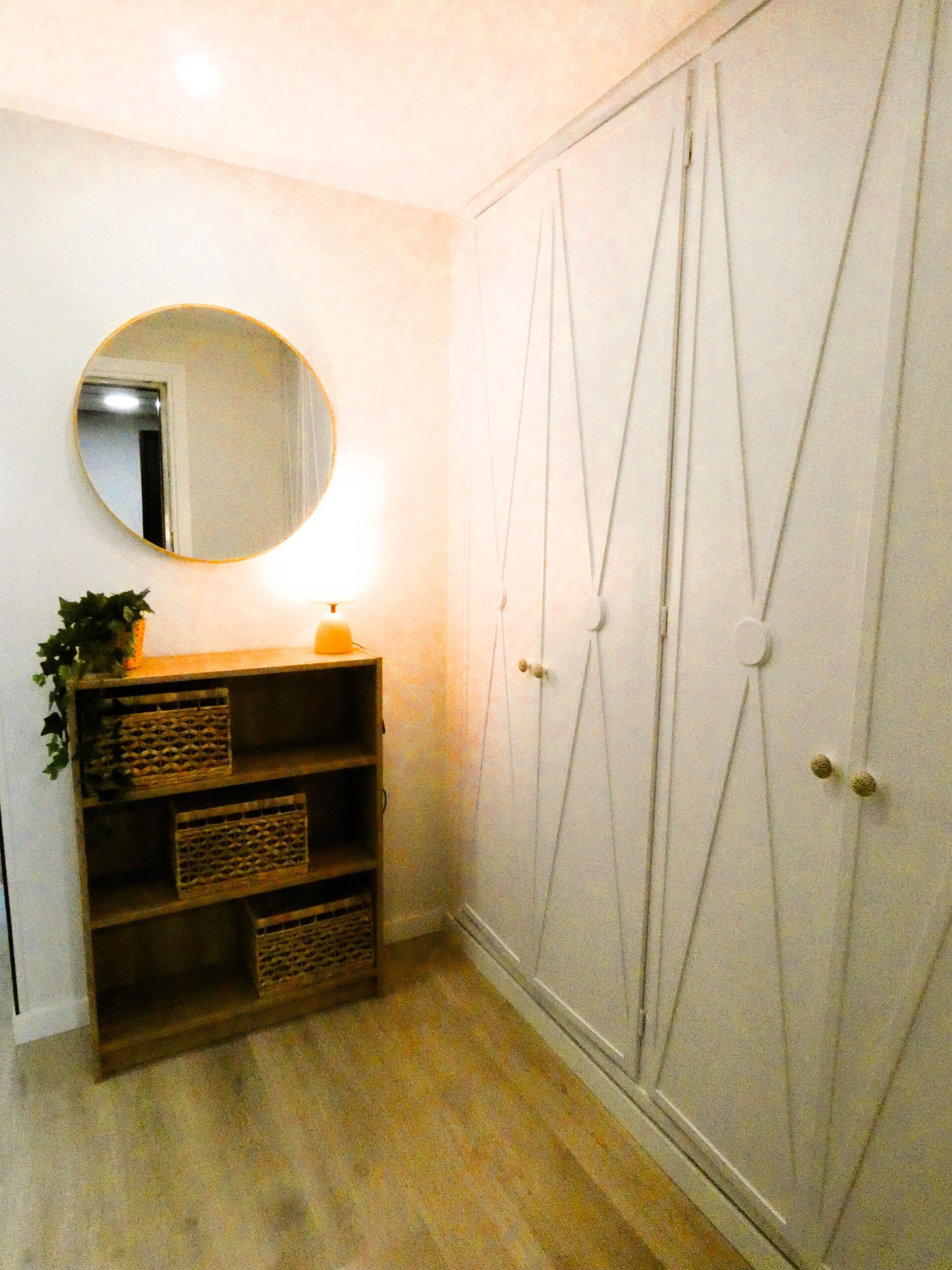 Una habitación con suelo de madera, un espejo redondo, una estantería con cestas y un armario con líneas verticales.