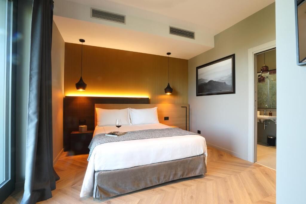 A modern hotel room with a bed, bedside tables, a nightstand, a painting on the wall, and a view of the bathroom.