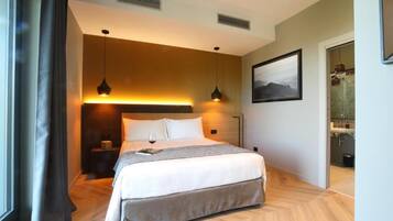 A modern hotel room with a bed, bedside tables, a nightstand, a painting on the wall, and a view of the bathroom.