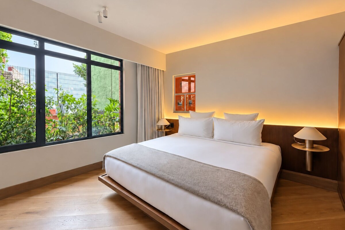 A hotel room with a large bed, a nightstand, a lamp, a window with a view of greenery, and a wall-mounted information board.