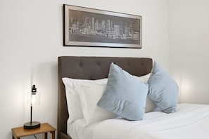 A neatly made bed with white and light blue pillows, a grey upholstered headboard, and a framed cityscape picture above. - Main Floor 2BR Retreat, 2 Parking, 1 Bath & Wi-Fi (Pickering)