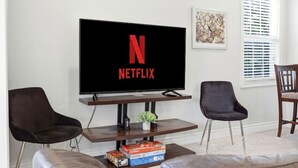 Ein Wohnzimmer mit einem Flachbildfernseher, der das Netflix-Logo anzeigt, zwei dunklen Sesseln, einem kleinen Tisch mit Gesellschaftsspielen und einem Fenster mit Jalousien.