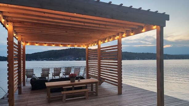 Eine Holzterrasse mit einer Pergola, Lichterketten und Gartenmöbeln mit Blick auf ein Gewässer.