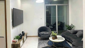 A modern living room with a flat-screen TV, a dark armchair, a sofa, a coffee table, and a large sliding glass door leading to a balcony. - Vinhomes central park  (Ho Chi Minh City)