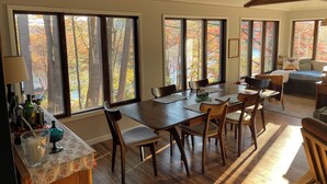 A dining area with a wooden table and chairs, large windows with autumn foliage views, and a cozy seating area with a sofa. - A conscious retreat overlooking the Delaware River (Beach Lake)