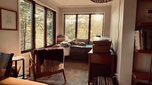 A cozy living room with a sofa, a chair, a small table, and a bookshelf. Large windows provide a view of trees outside. - A conscious retreat overlooking the Delaware River (Beach Lake)