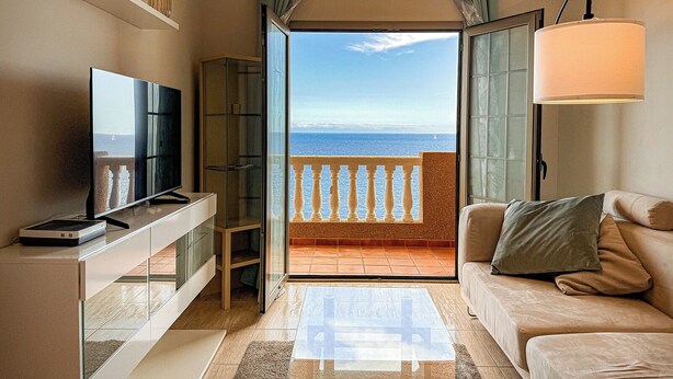 Una sala de estar con televisor, sofá, una mesita de centro de cristal y un balcón con vistas al mar.