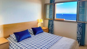 Un dormitorio con cama, mesita de noche, lámpara, ventana y vistas al mar, además de cortinas.