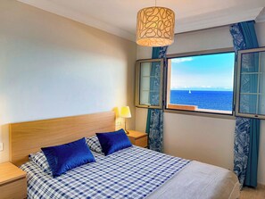 A bedroom with a bed, a nightstand, a lamp, a window with a view of the sea, and curtains. - Apartment with breathtaking view (Porís de Abona)