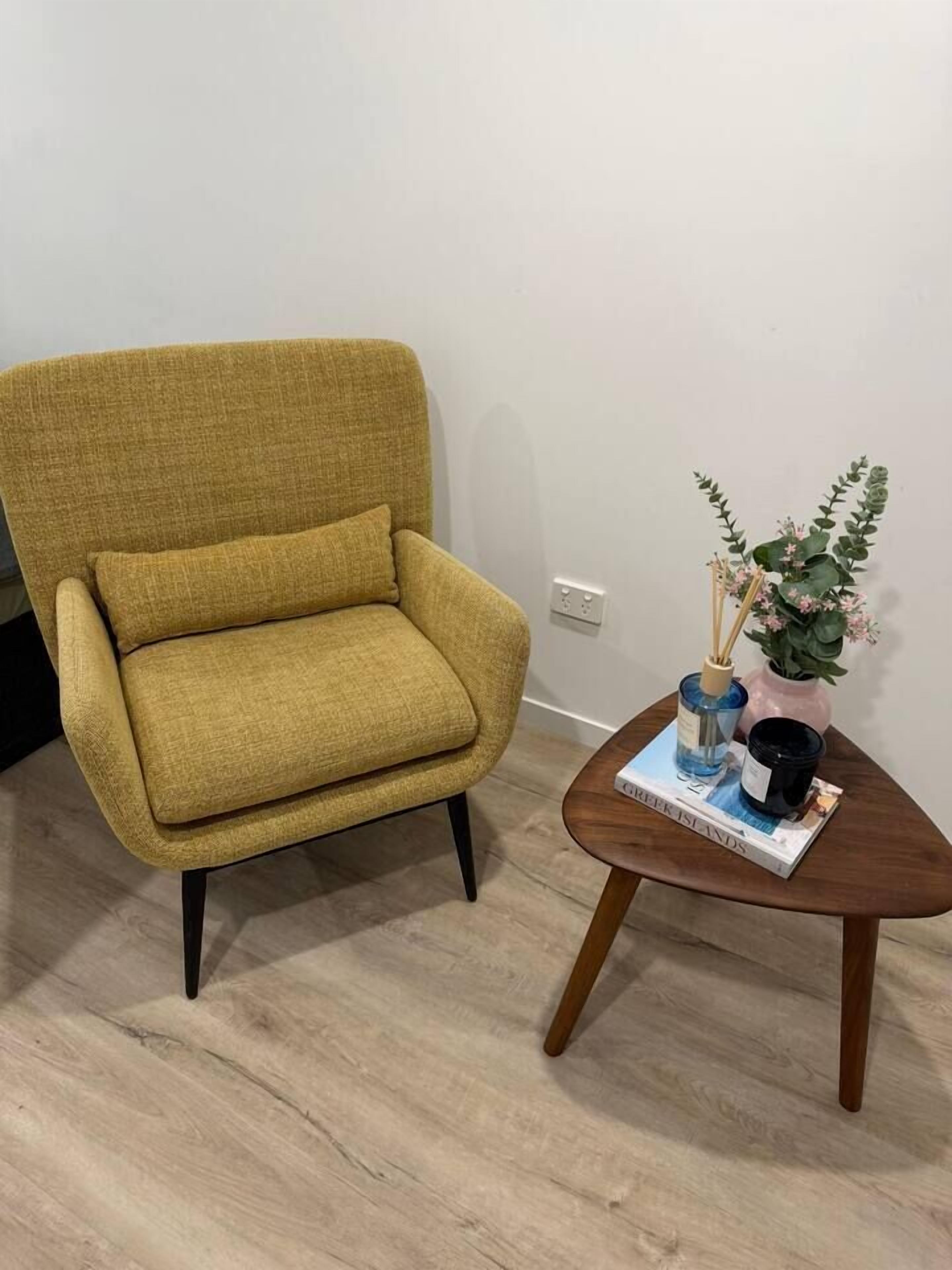 A mustard-colored armchair with black legs, a small wooden side table with a book, a blue bottle, and a pink vase with green foliage.