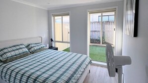 A bedroom with a bed, a standing fan, and a window with a view of a backyard.
