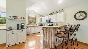 A kitchen with white cabinets, a wooden island, bar stools, a clock, and a coffee bar sign. - 5BR Outer Banks Retreat ✦ Pool ✦ Spa ✦ Private Beach Access Prkg ✦ Dog-Friendly (Southern Shores)