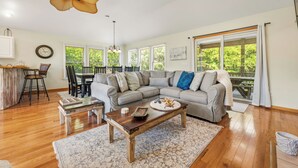 A spacious living room with a gray sectional sofa, a wooden coffee table, a dining area with a table and chairs, and large windows offering a view of greenery. - 5BR Outer Banks Retreat ✦ Pool ✦ Spa ✦ Private Beach Access Prkg ✦ Dog-Friendly (Southern Shores)