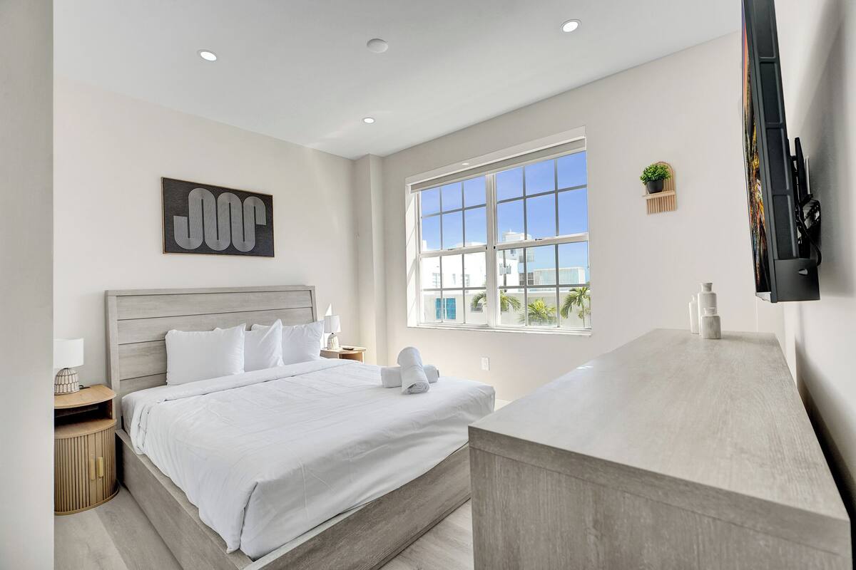 A modern hotel room with a large bed, a TV, a window with a city view, and a minimalist decor.