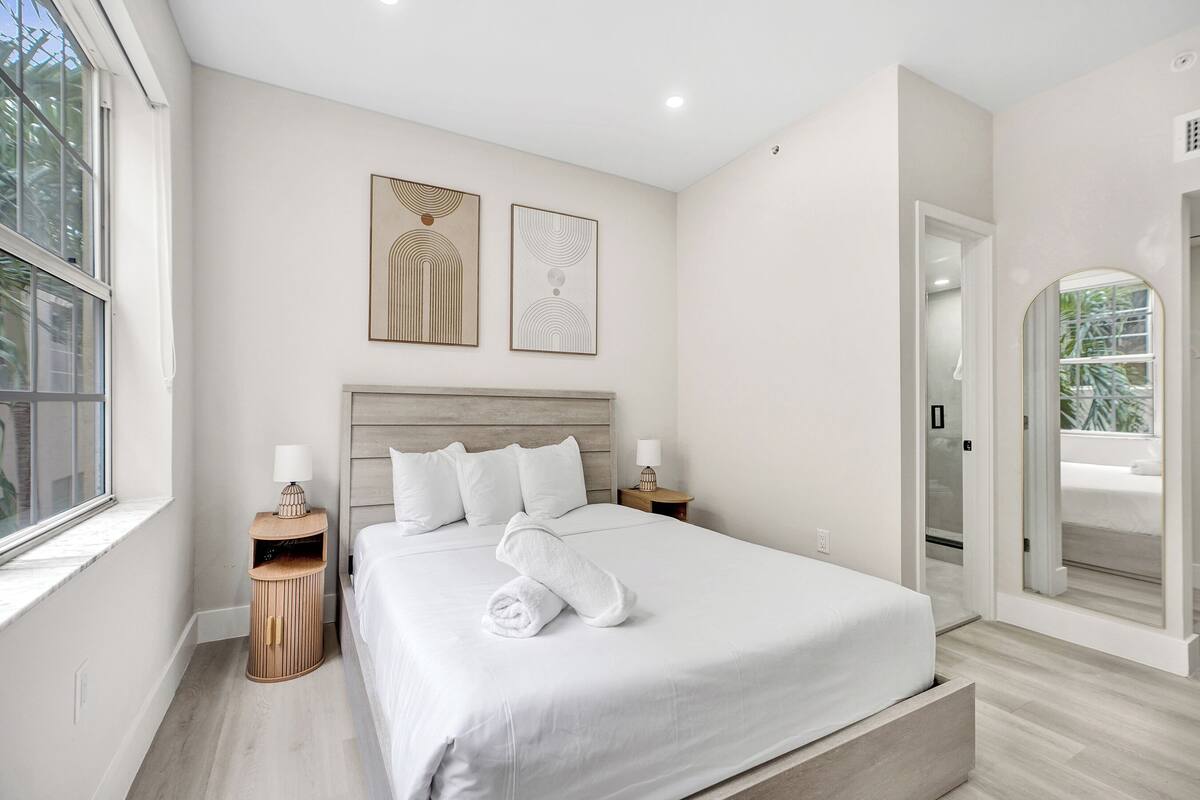 A modern hotel room with a large bed, a bedside table with a lamp, a small side table, and a view of the outside through a window.