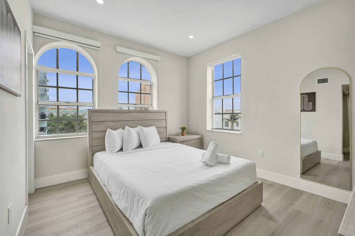 A bedroom with a large bed, two windows, a nightstand, a mirror, and a view of buildings outside.