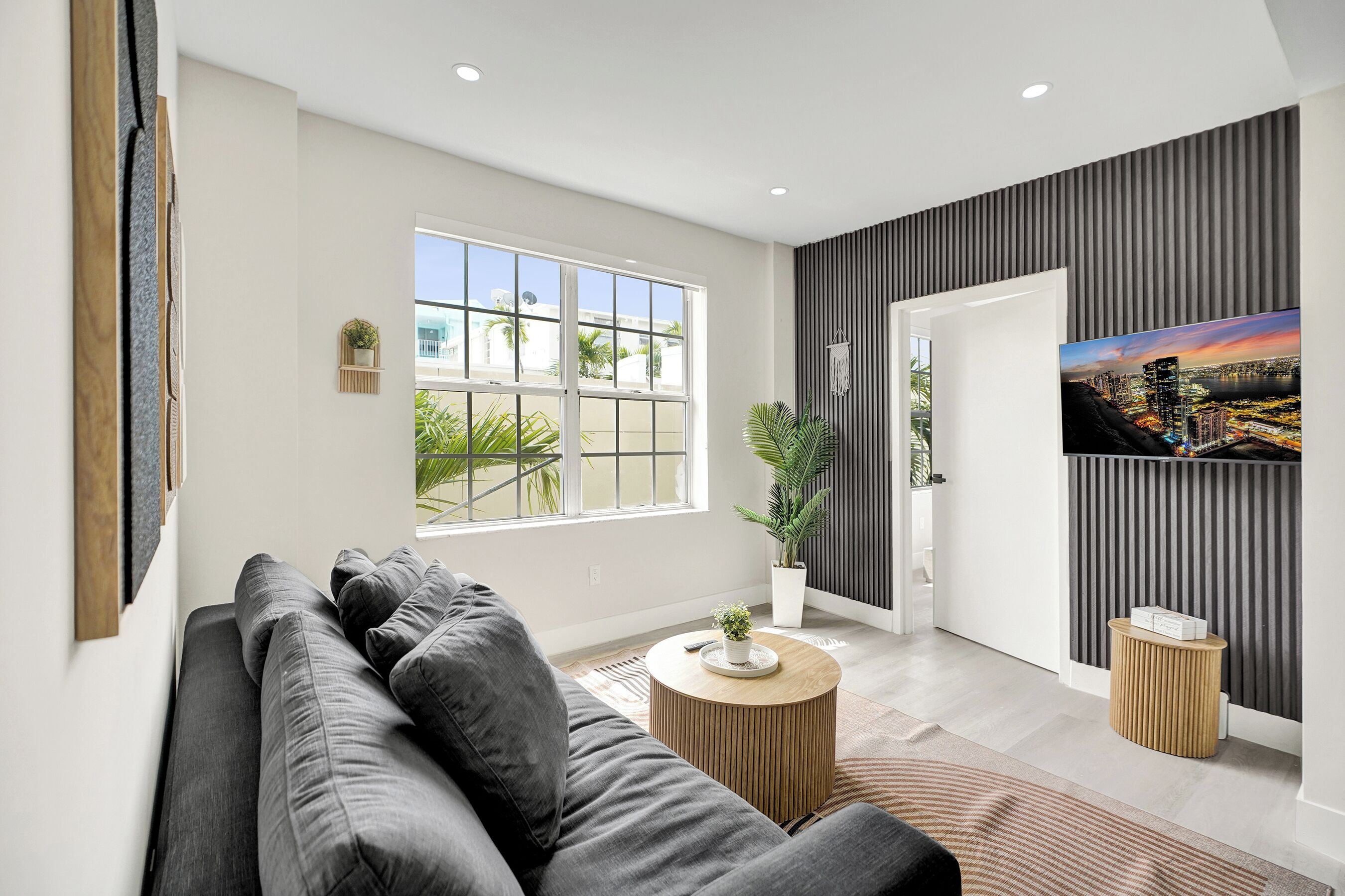 Een moderne woonkamer met een grijze bank, een ronde houten salontafel, een kamerplant en een stadslandschap op de muur.